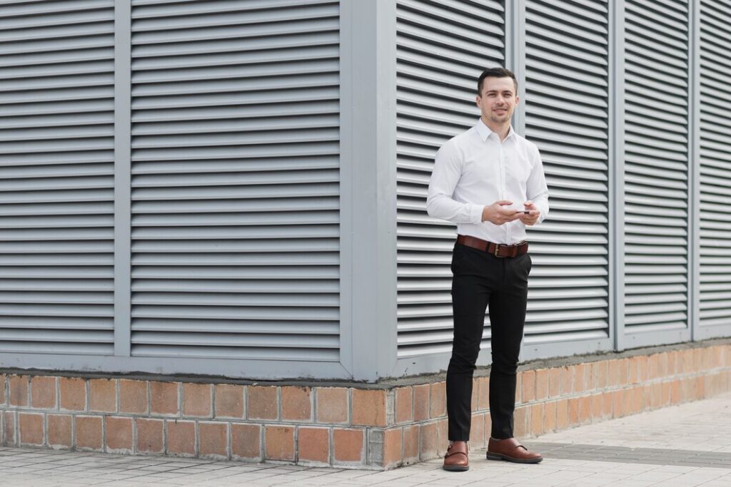 Man in a white shirt and black trousers standing against a modern building wall.