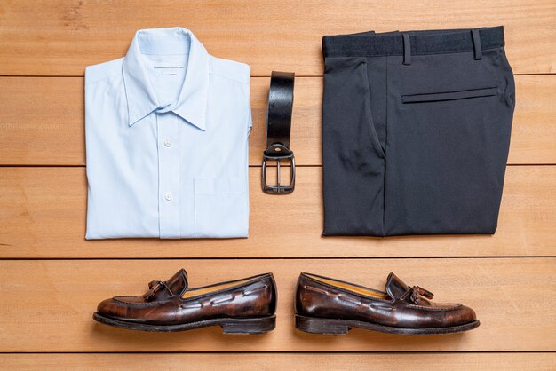 Flat lay of a folded blue dress shirt, black trousers, leather belt, and brown loafers arranged neatly on a wooden surface.